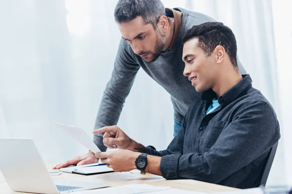 Sonriente joven empresario señalando con el dedo a los papeles mientras trabaja con su colega en la oficina - foto de stock