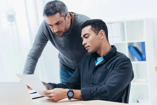 Bearded mentor looking at young colleague working with papers in office — Stock Photo