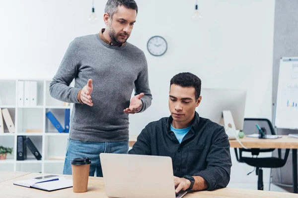 Bärtiger Geschäftsmann gestikuliert mit den Händen und schaut junge Kollegin an, die mit Laptop arbeitet — Stockfoto