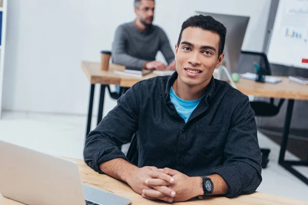 Handsome young businessman sitting at workplace and smiling at camera — Stock Photo