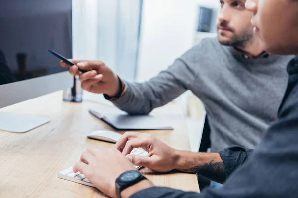 Recortado disparo de compañeros de trabajo escribiendo en el teclado y apuntando a la pantalla mientras se trabaja con la computadora de escritorio juntos - foto de stock