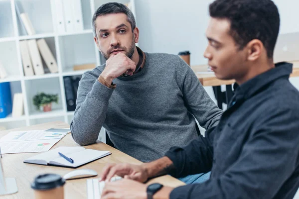 Mentor sérieux barbu entreprise regardant jeune collègue en utilisant un ordinateur de bureau sur le lieu de travail — Photo de stock