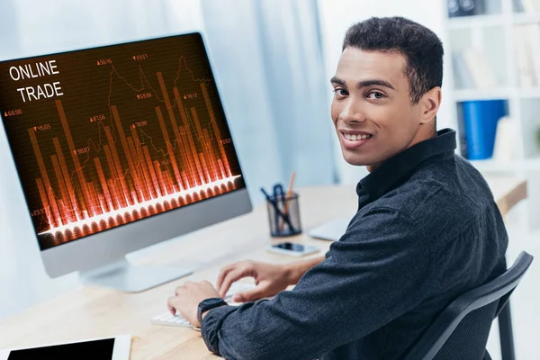 Junge gemischte Rasse Geschäftsmann verwendet Desktop-Computer mit Online-Handelstabellen und lächelt in die Kamera — Stockfoto