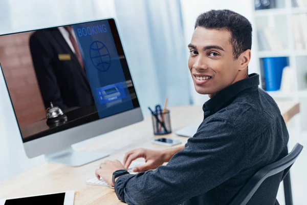 Young mixed race businessman using desktop computer with booking website and smiling at camera — Stock Photo