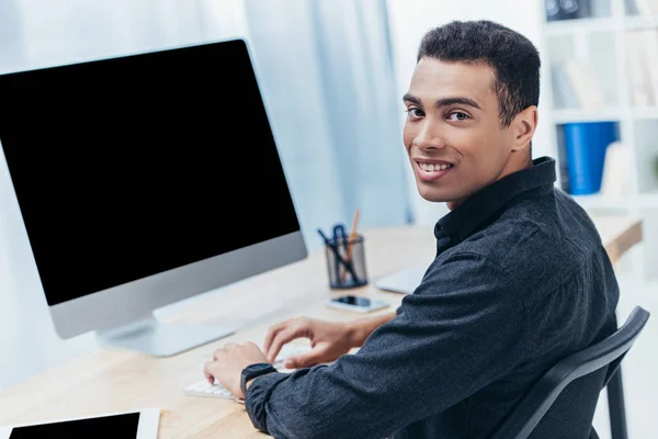 Joven hombre de negocios de raza mixta utilizando el ordenador de sobremesa con pantalla en blanco y sonriendo a la cámara - foto de stock