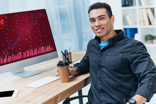 Young businessman with coffee to go working with desktop computer with business charts and smiling at camera — Stock Photo