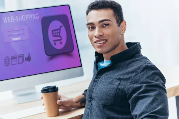 Jeune homme d'affaires avec café pour aller travailler avec ordinateur de bureau avec site de shopping et sourire à la caméra — Photo de stock