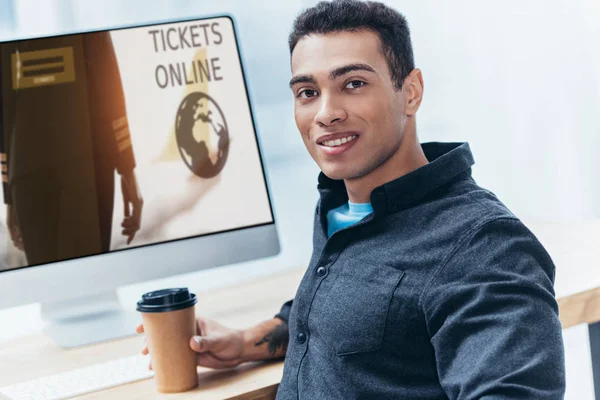 Jovem empresário com café para ir trabalhar com computador desktop com bilhetes site on-line e sorrindo para a câmera — Fotografia de Stock