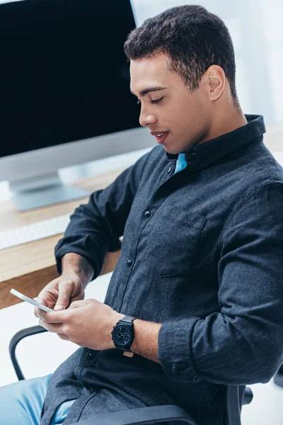 Hochwinkelaufnahme eines lächelnden jungen Geschäftsmannes, der im Büro sitzt und sein Smartphone benutzt — Stockfoto