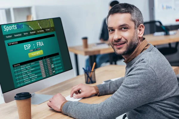 Schöner bärtiger Geschäftsmann arbeitet mit Desktop-Computer mit Sportwetten-Website und lächelt im Büro in die Kamera — Stockfoto