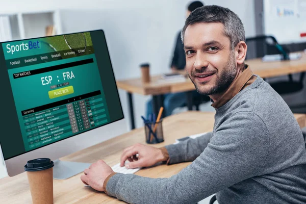 Beau barbu homme d'affaires travaillant avec ordinateur de bureau avec site de paris sportifs et souriant à la caméra dans le bureau — Photo de stock