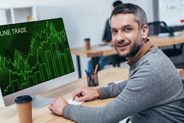 Handsome bearded businessman working with desktop computer with online trade graphs and smiling at camera in office — Stock Photo