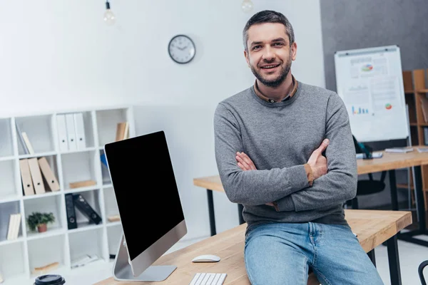 Schöner bärtiger Geschäftsmann sitzt mit verschränkten Armen auf dem Schreibtisch und lächelt in die Kamera im Büro — Stockfoto