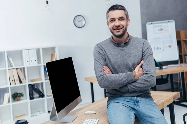 Bonito empresário barbudo sentado com os braços cruzados na mesa e sorrindo para a câmera no escritório — Fotografia de Stock