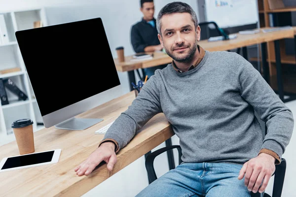 Schöner Mann blickt in die Kamera, während er mit Desktop-Computer und digitalem Tablet im Büro am Tisch sitzt — Stockfoto