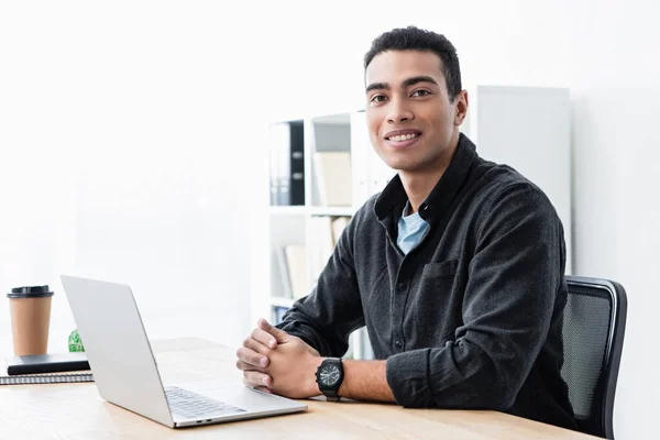 Fröhliche junge gemischte Rasse Geschäftsmann sitzt am Tisch mit Laptop und lächelt in die Kamera im Büro — Stockfoto