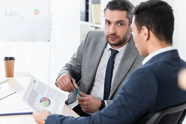 Serious bearded businessman looking at young male colleague holding document with charts in office — Stock Photo