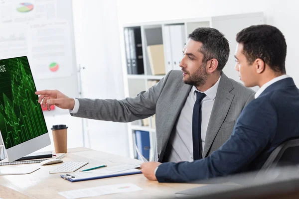 Bearded businessman showing desktop computer with trading graphs on screen to young colleague at workplace — Stock Photo