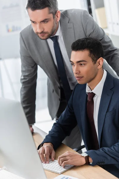 Mentor de negocios barbudo mirando a un joven colega utilizando la computadora de escritorio en el lugar de trabajo - foto de stock