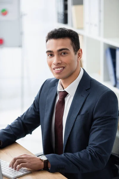 Schöner junger Geschäftsmann, der mit Desktop-Computer arbeitet und im Büro in die Kamera lächelt — Stockfoto