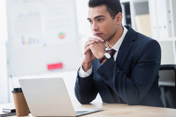 Confiante jovem empresário sentado à mesa com laptop e olhando para longe no escritório — Stock Photo