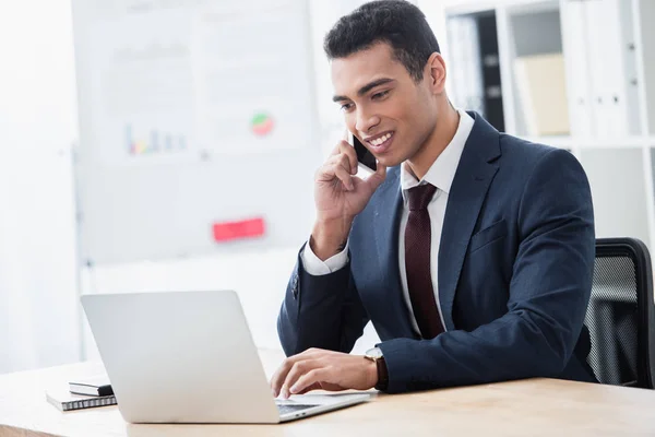 Sonriente joven hombre de negocios hablando por teléfono inteligente y trabajando con el ordenador portátil en la oficina - foto de stock