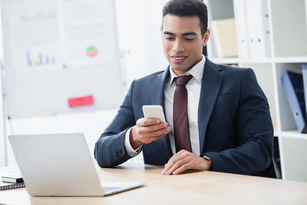 Sorridente jovem empresário segurando smartphone e trabalhando com laptop no escritório — Fotografia de Stock