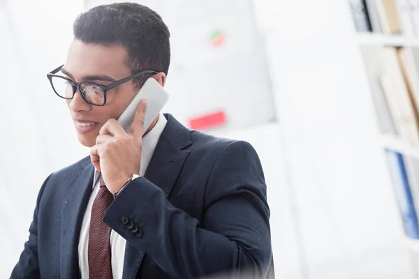 Smiling young businessman in eyeglasses talking by smartphone and looking away in office — Stock Photo