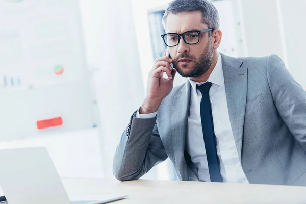 Uomo d'affari barbuto concentrato negli occhiali che parla con lo smartphone e lavora con il computer portatile in ufficio — Foto stock