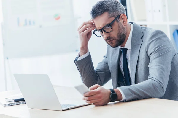 Hombre de negocios concentrado en gafas de vista usando smartphone y trabajando con laptop en oficina - foto de stock