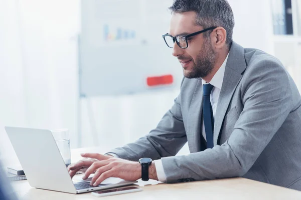 Усміхнений бородатий бізнесмен в окулярах використовує ноутбук на робочому місці — стокове фото