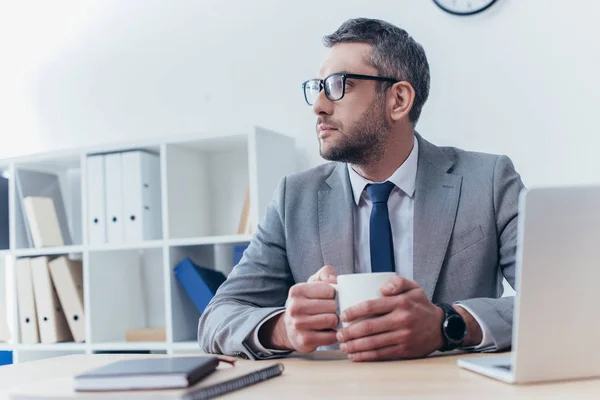Серьезный бизнесмен в очках держит чашку кофе и смотрит в сторону, сидя на рабочем месте — стоковое фото