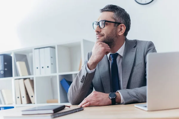 Pensive businessman in eyeglasses sitting at workplace and looking away — Stock Photo