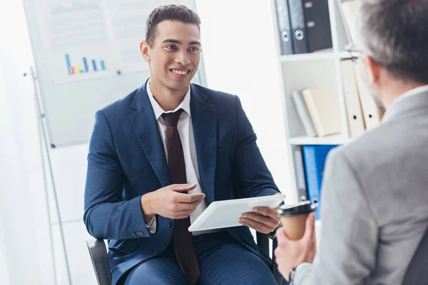 Recortado disparo de sonriente joven hombre de negocios utilizando tableta digital y mirando colega masculino con taza de papel - foto de stock