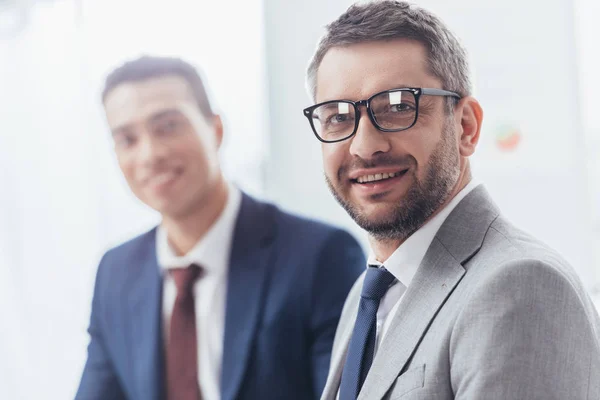 Handsome bearded businessman in eyeglasses smiling at camera, young colleague standing behind — Stock Photo