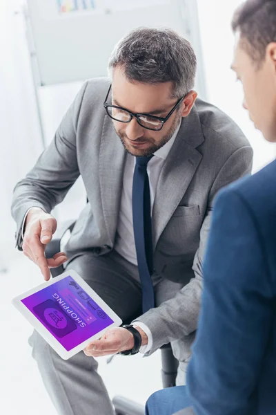 Cropped shot of businessman in eyeglasses showing digital tablet with online shopping app to male colleague in office — Stock Photo