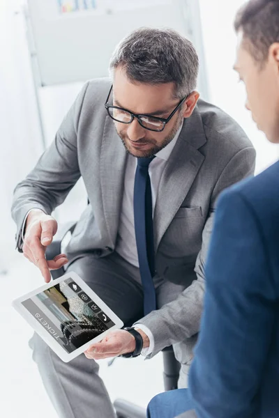 Cropped shot of businessman in eyeglasses showing digital tablet with online tickets app to male colleague in office — Stock Photo