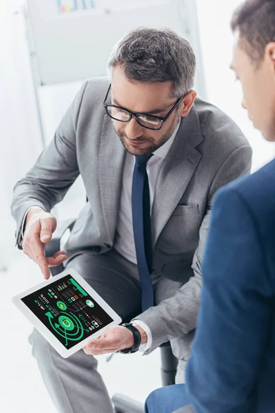 Cropped shot of businessman in eyeglasses showing digital tablet with business charts on screen to male colleague in office — Stock Photo