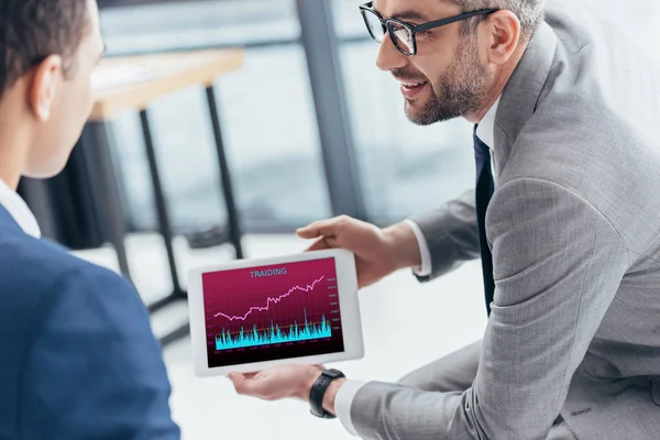 Cropped shot of businessman in eyeglasses showing digital tablet with trading graphs on screen to male colleague in office — Stock Photo