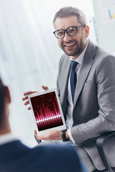 Smiling businessman in eyeglasses holding digital tablet with business charts on screen — Stock Photo