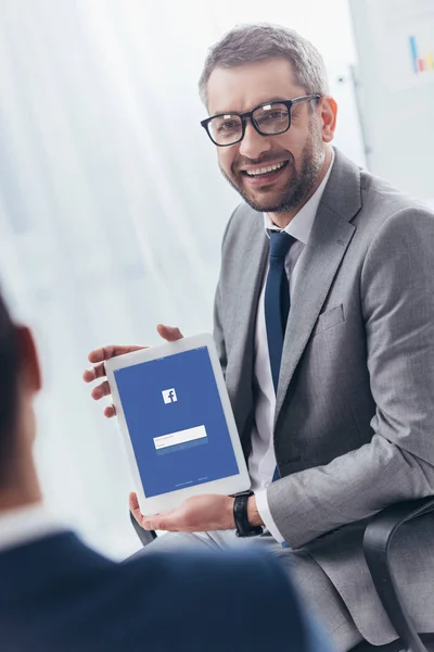 Усміхаючись бізнесмен в окулярів проведення Цифрова табличка з facebook app на екрані — стокове фото