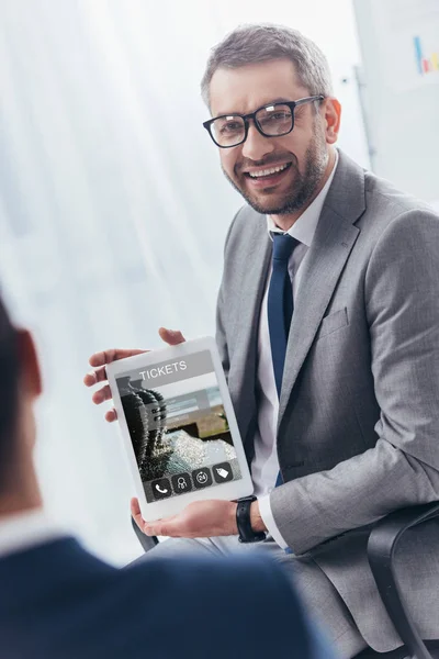 Улыбающийся бизнесмен в очках, держащий цифровой планшет с онлайн билетами на экране — стоковое фото