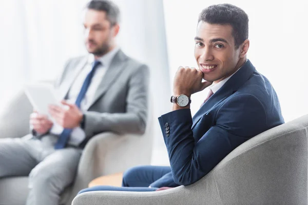 Handsome young businessman sitting and smiling at camera while male colleague using digital tablet behind — Stock Photo