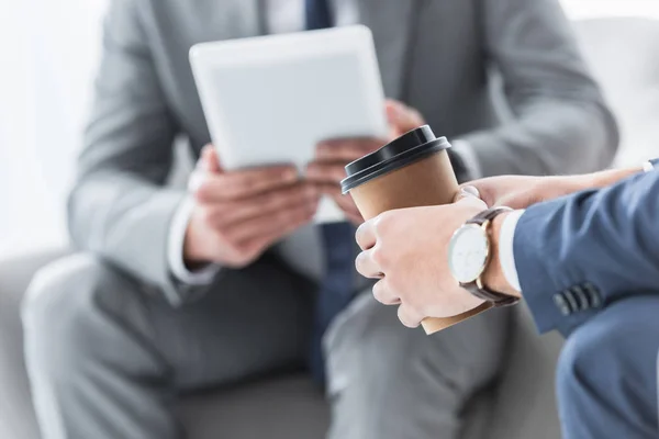 Recortado disparo de empresario sosteniendo café para ir y colega utilizando tableta digital detrás - foto de stock