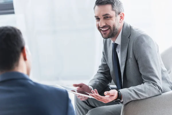 Sonriente joven empresario sosteniendo tableta digital y mirando a su colega masculino en primer plano - foto de stock