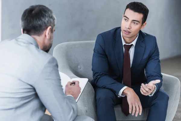 Jungunternehmer spricht und schaut männlichen Kollegen zu, der sitzt und Notizen in Notizbuch macht — Stockfoto