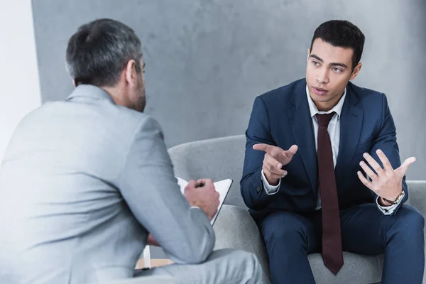 Joven hombre de negocios hablando y mirando mentor de negocios sentado y tomando notas en el cuaderno - foto de stock