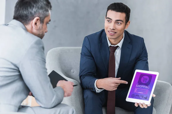 Smiling young businessman pointing at digital tablet with shopping app on screen and looking at male colleague during conversation — Stock Photo