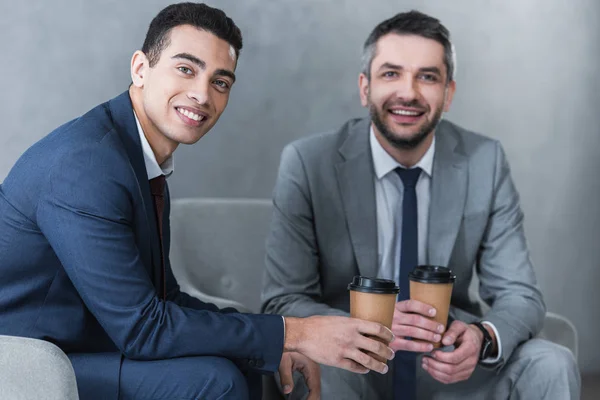 Uomini d'affari professionisti che tengono il caffè per andare e sorridere alla macchina fotografica mentre seduti insieme — Foto stock