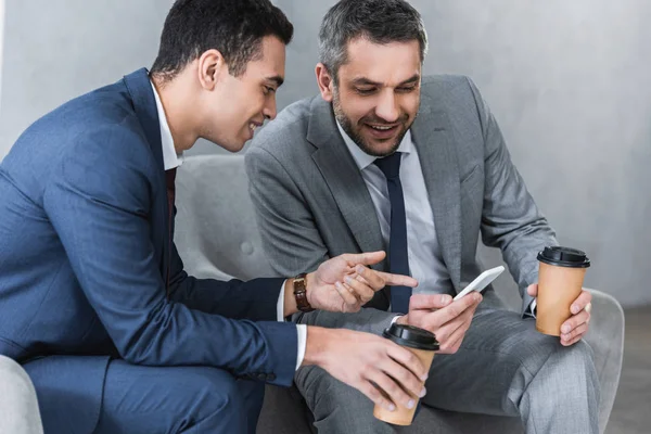 Homens de negócios sorrindo segurando café para ir e usar o smartphone juntos — Fotografia de Stock